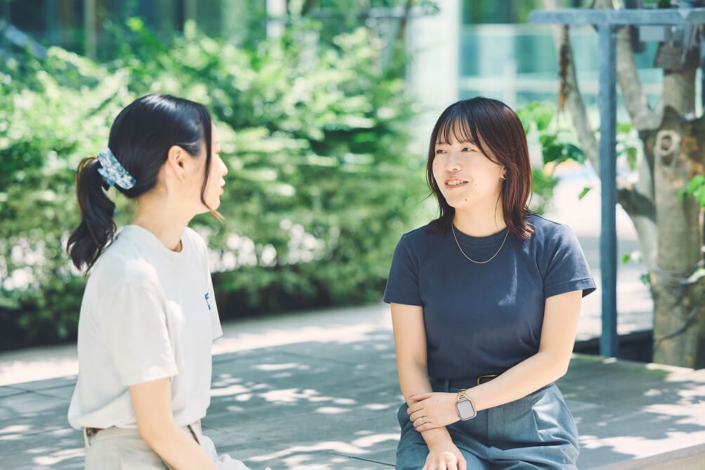 屋外でベンチに座り向き合いながら楽しそうに話している様子の新留
